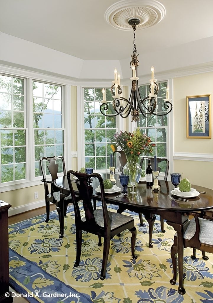 The dining room offers an oval dining set, an ornate chandelier, a large floral rug, and a bay window.