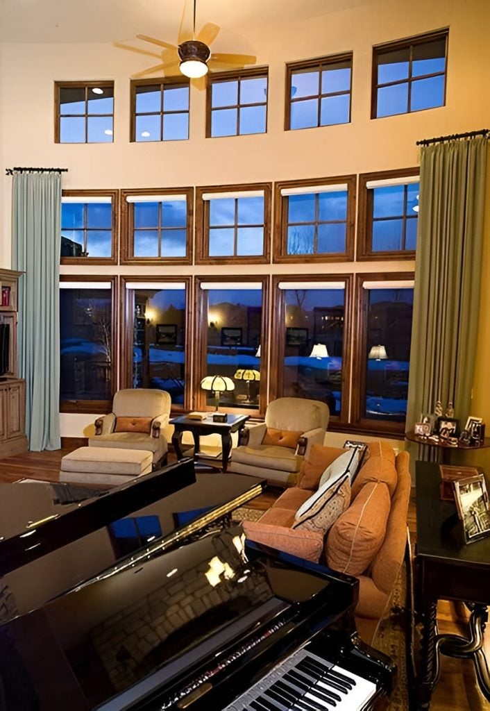Living room with comfy fabric seats, a baby grand piano, and lots of wooden framed windows dressed in green draperies.
