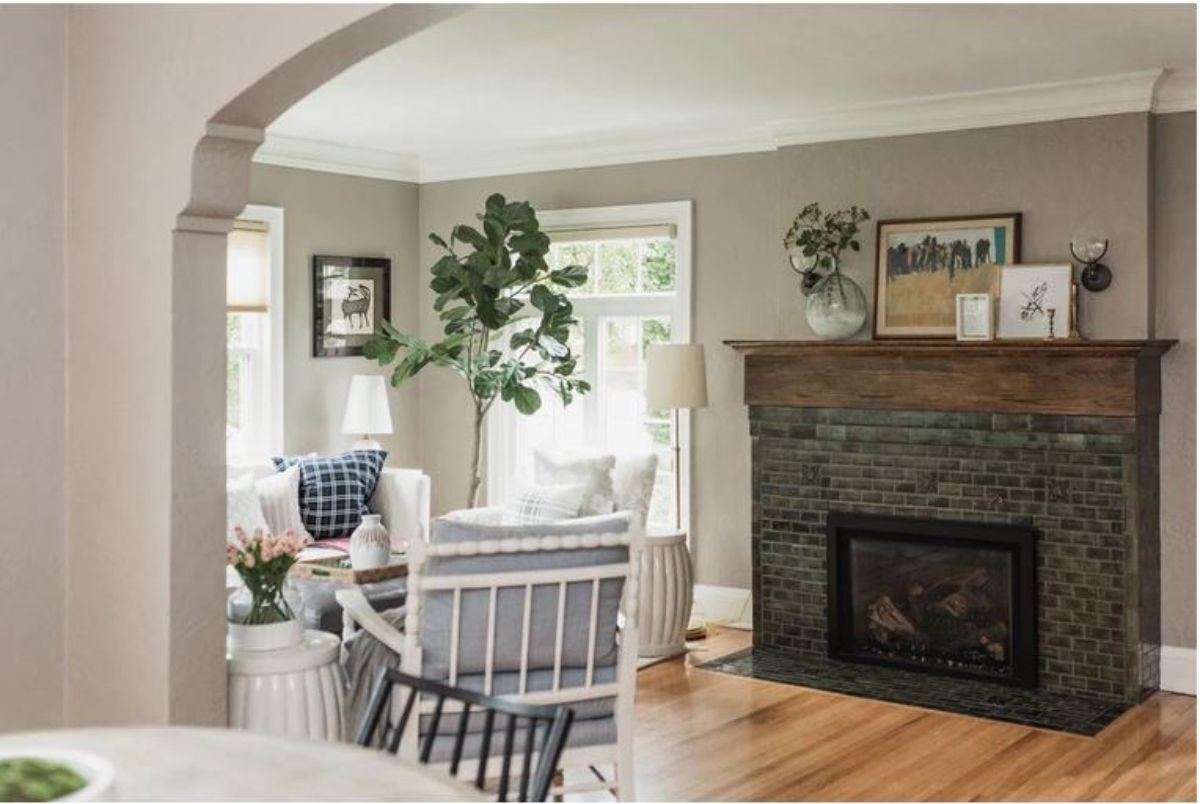 A fireplace with art frames and a plant in a vase, chairs beside the window, and an indoor plant.