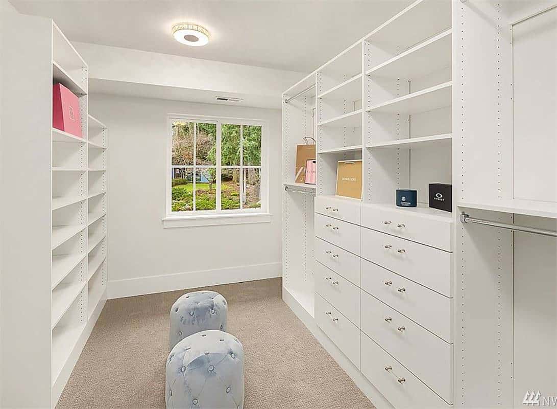 Primary walk-in closet with white built-ins, tufted stools, and a white-framed window that invites natural light in.