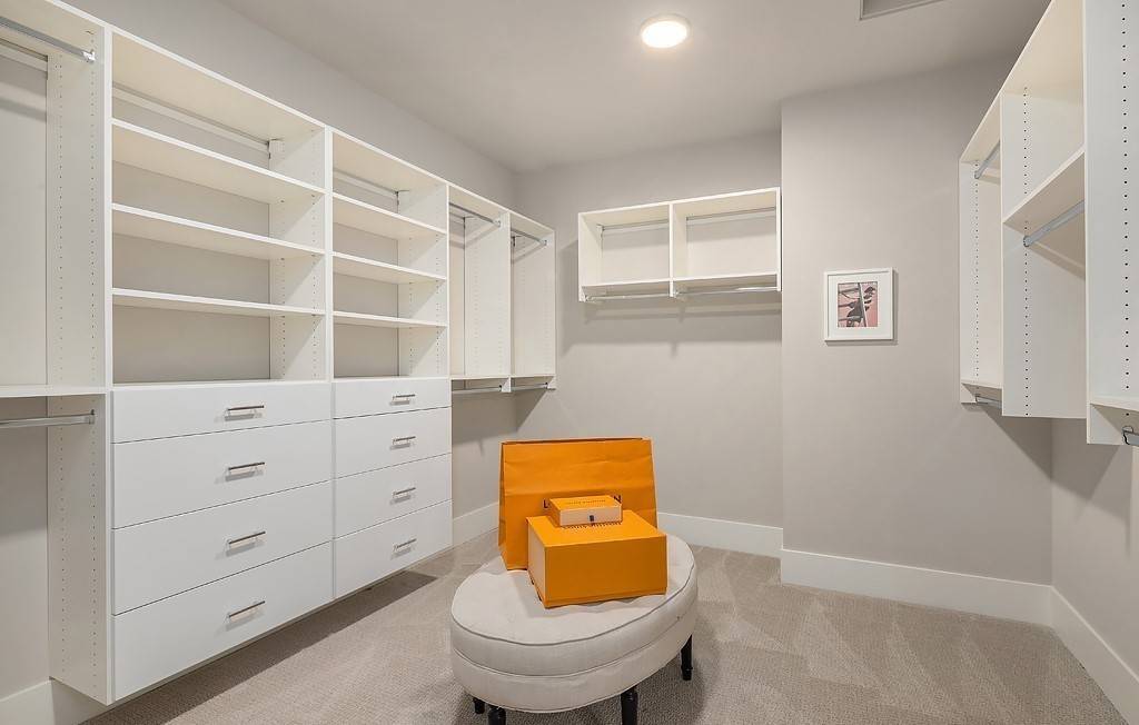 Primary walk-in closet with white built-ins and a tufted ottoman over the carpeted floor.