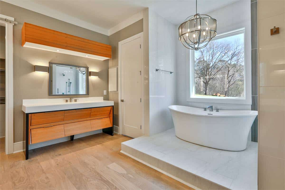 Primary bathroom with a large vanity and a freestanding tub illuminated by a chrome chandelier and a picture window.