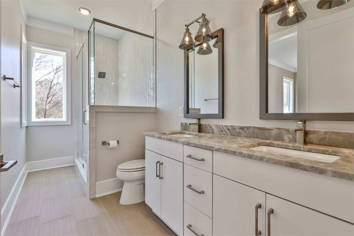 Shared bathroom with a dual sink vanity, a toilet, and a walk-in shower brightened by corner windows.