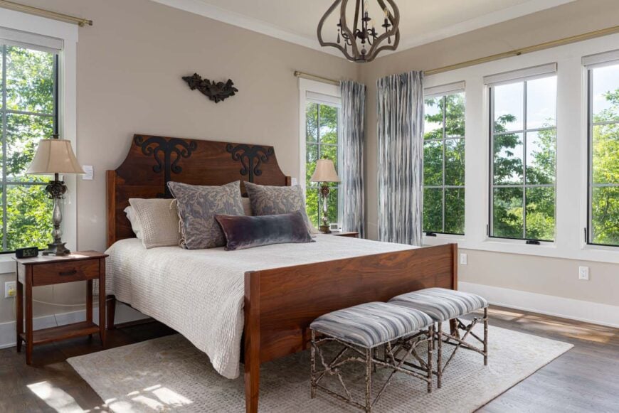 Bed with a wooden frame and white comforter in a bedroom positioned between two bedside lamps and surrounded by windows