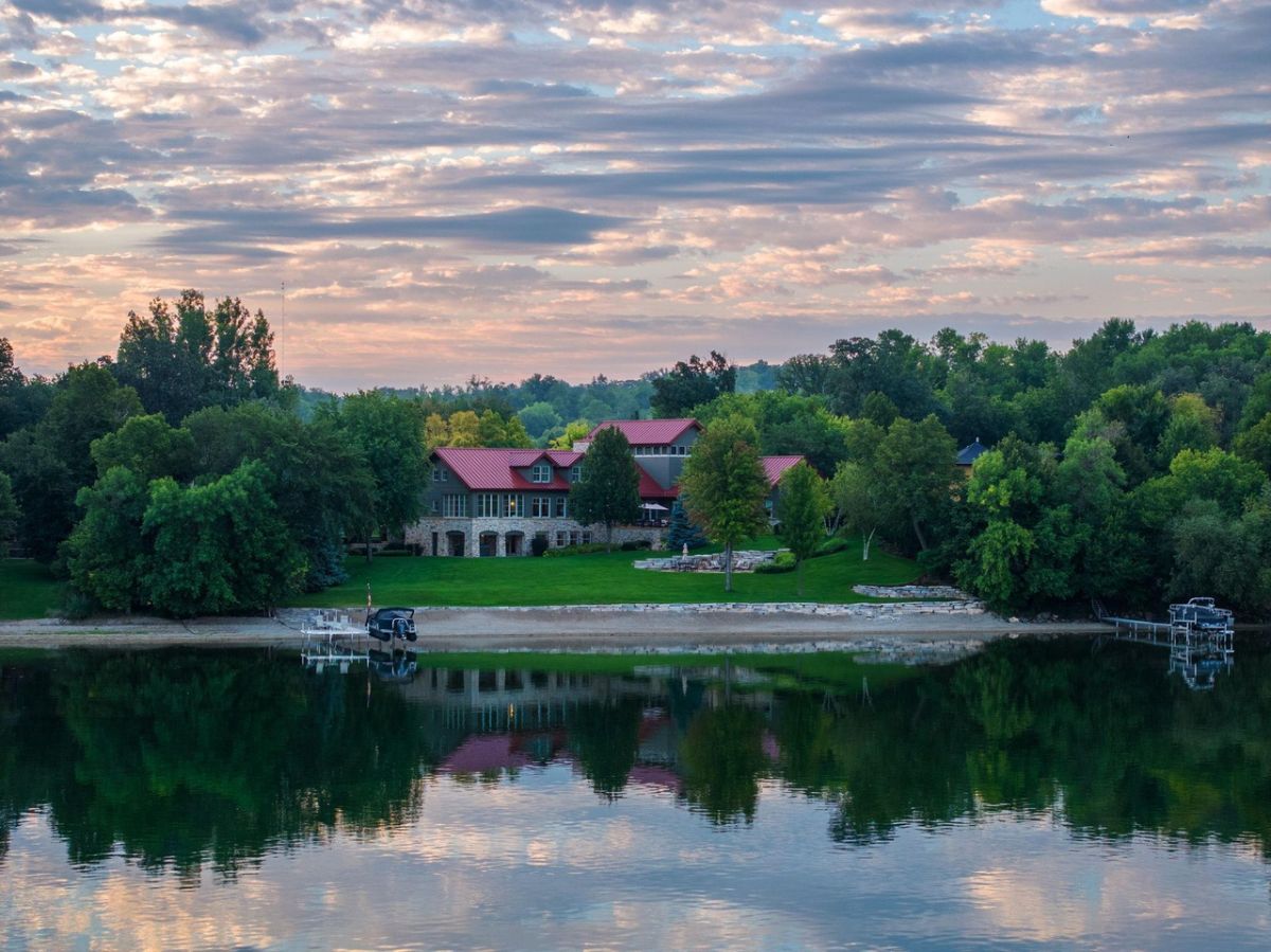 Aerial rear view of the house with a a large green lawn, a lake shore and trees in the background.