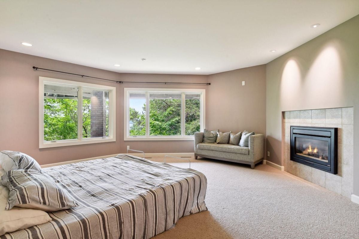 Bedroom with carpet flooring, a large bed, couch, fireplace and glass windows.
