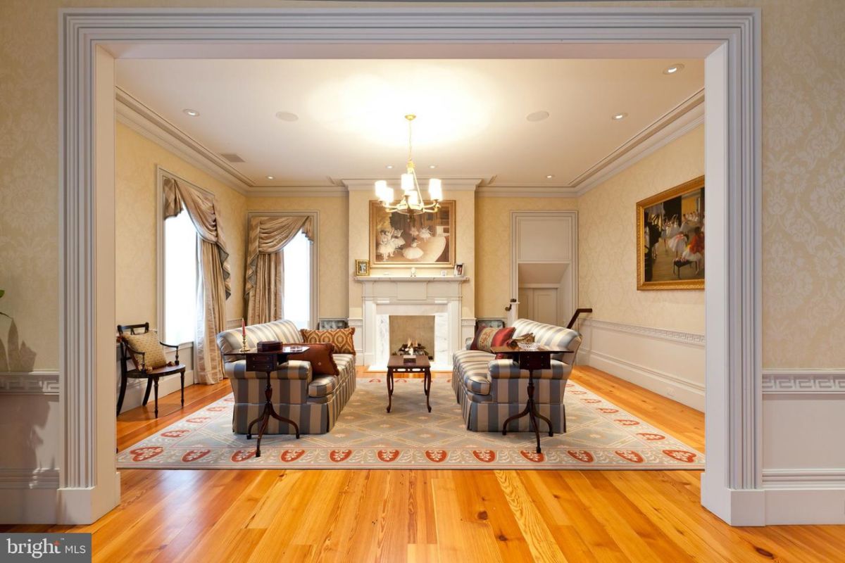 Living room with wooden floors, couches, chandelier, a large rug large glass windows and a fireplace.