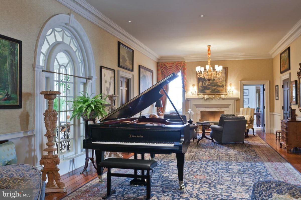 Music room with wooden floors, couches, chandelier, a large rug, large glass windows and a fireplace.