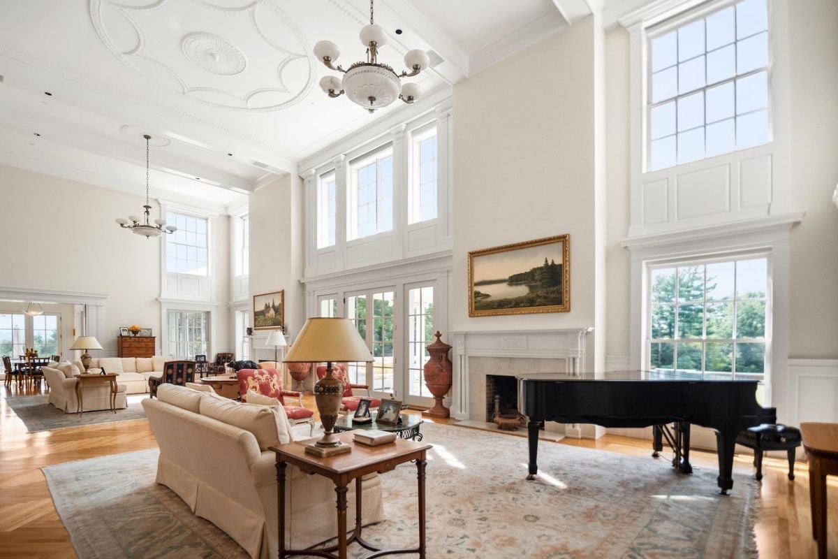 Living room with couches, high ceilings, chandeliers, piano, glass windows and a fireplace.