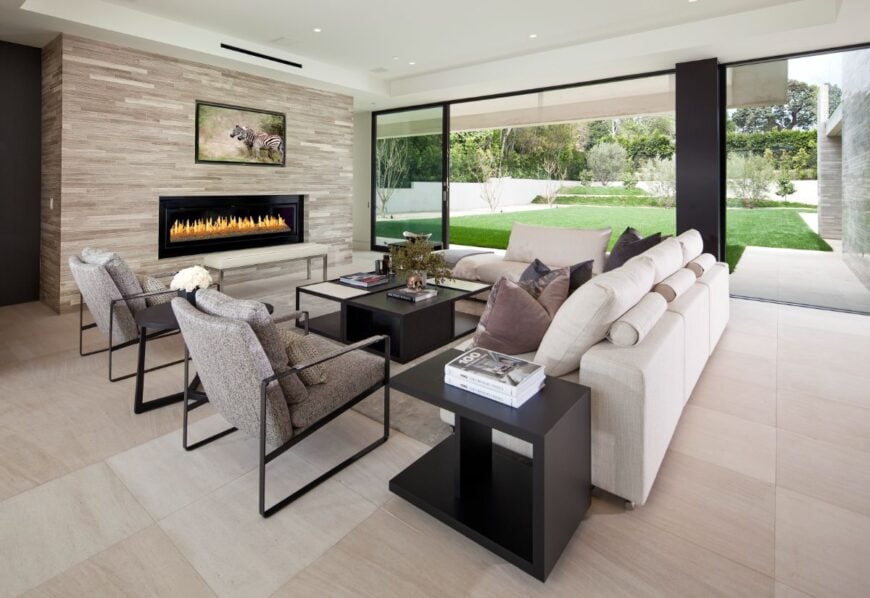Lower level family room wraps large white L-shaped sectional and twin metal frame armchairs around a cubic black coffee table, in front of massive gas fireplace surround. Floor to ceiling glass allows for expansive views over back yard.