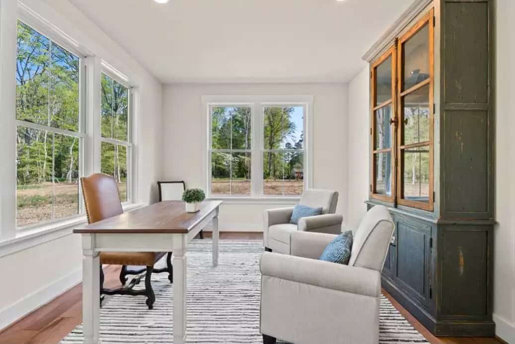 Home office with a wooden desk, gray armchairs, a display cabinet, and expansive backyard views.