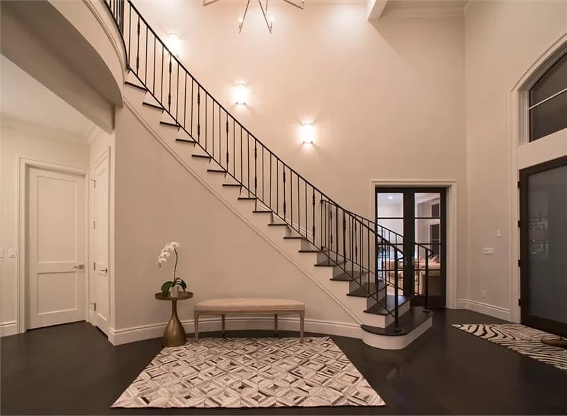 The foyer includes a flower pedestal and a cushioned bench against the curved staircase.
