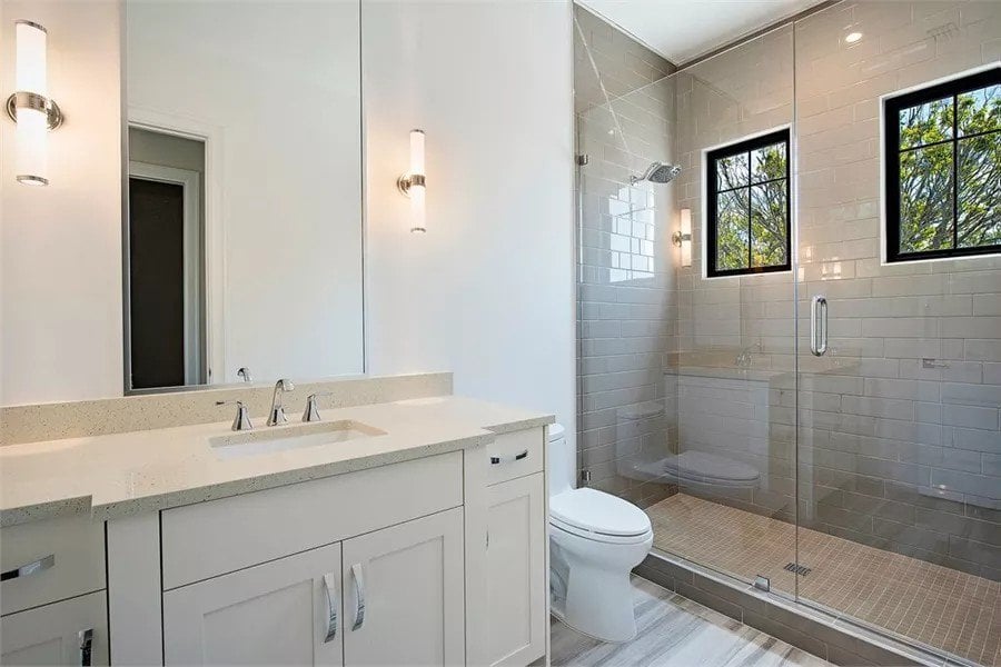 Bathroom with a sink vanity, a toilet, and a walk-in shower brightened by black framed windows.