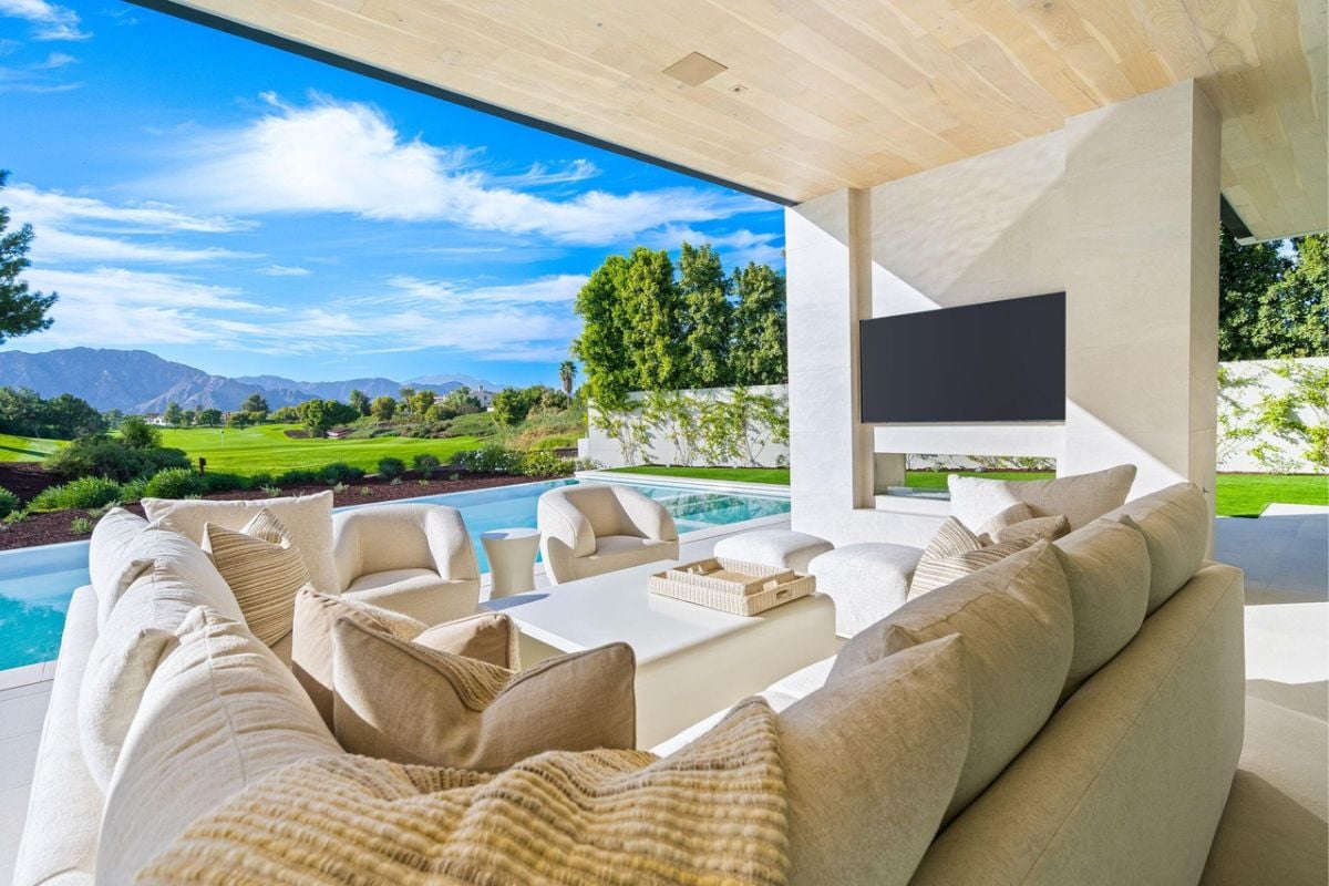 A covered patio furnished with white couches, pillows, a center table, and featuring a TV.