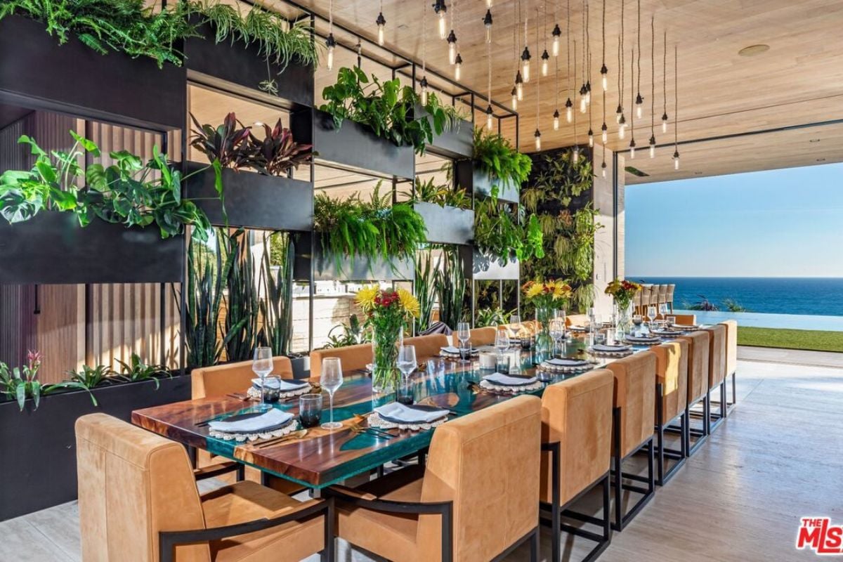 A dining room with plants, lighting, and a long table surrounded by chairs.