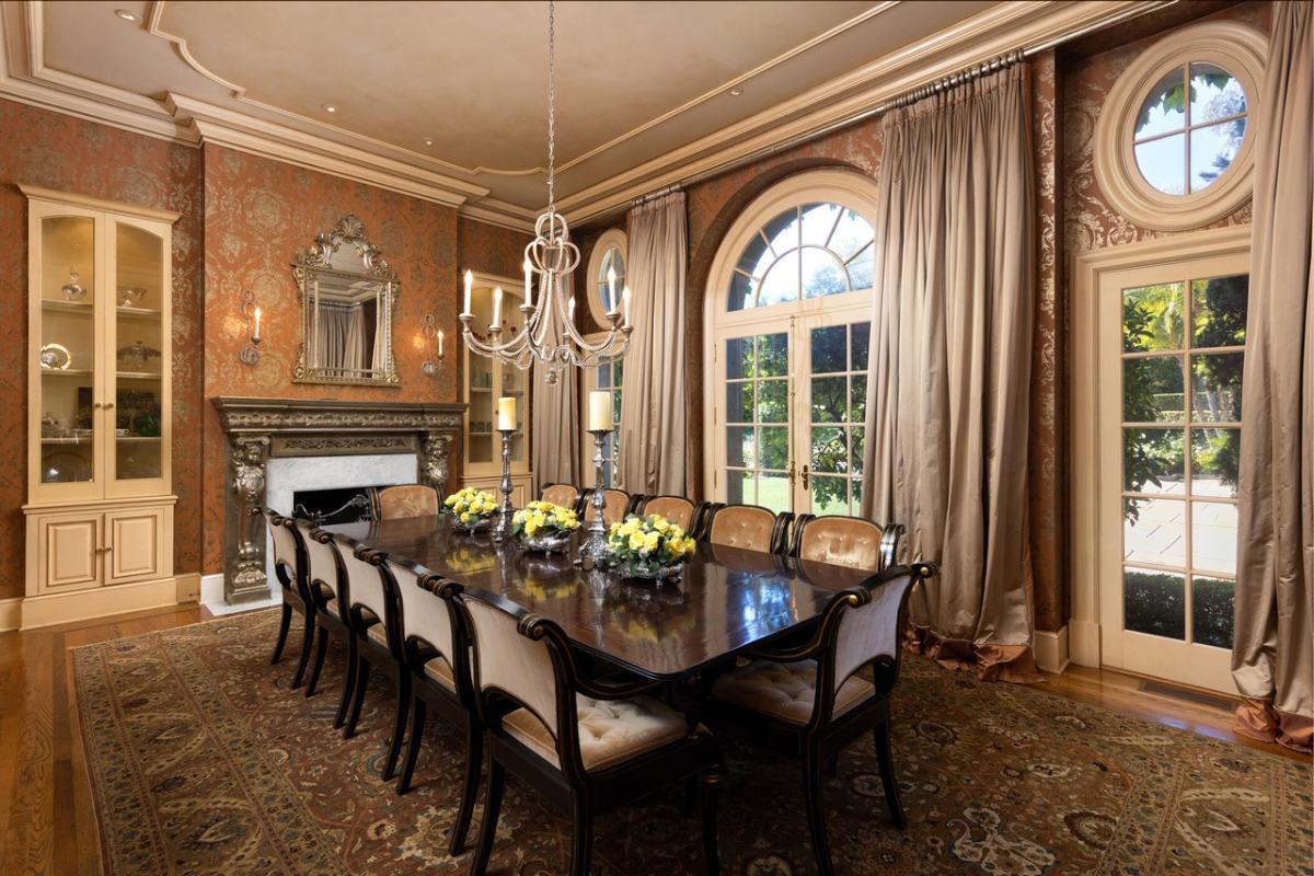 A dining room table with chairs around it. The room features a wall, window, floor, ceiling, curtain, and chandelier.