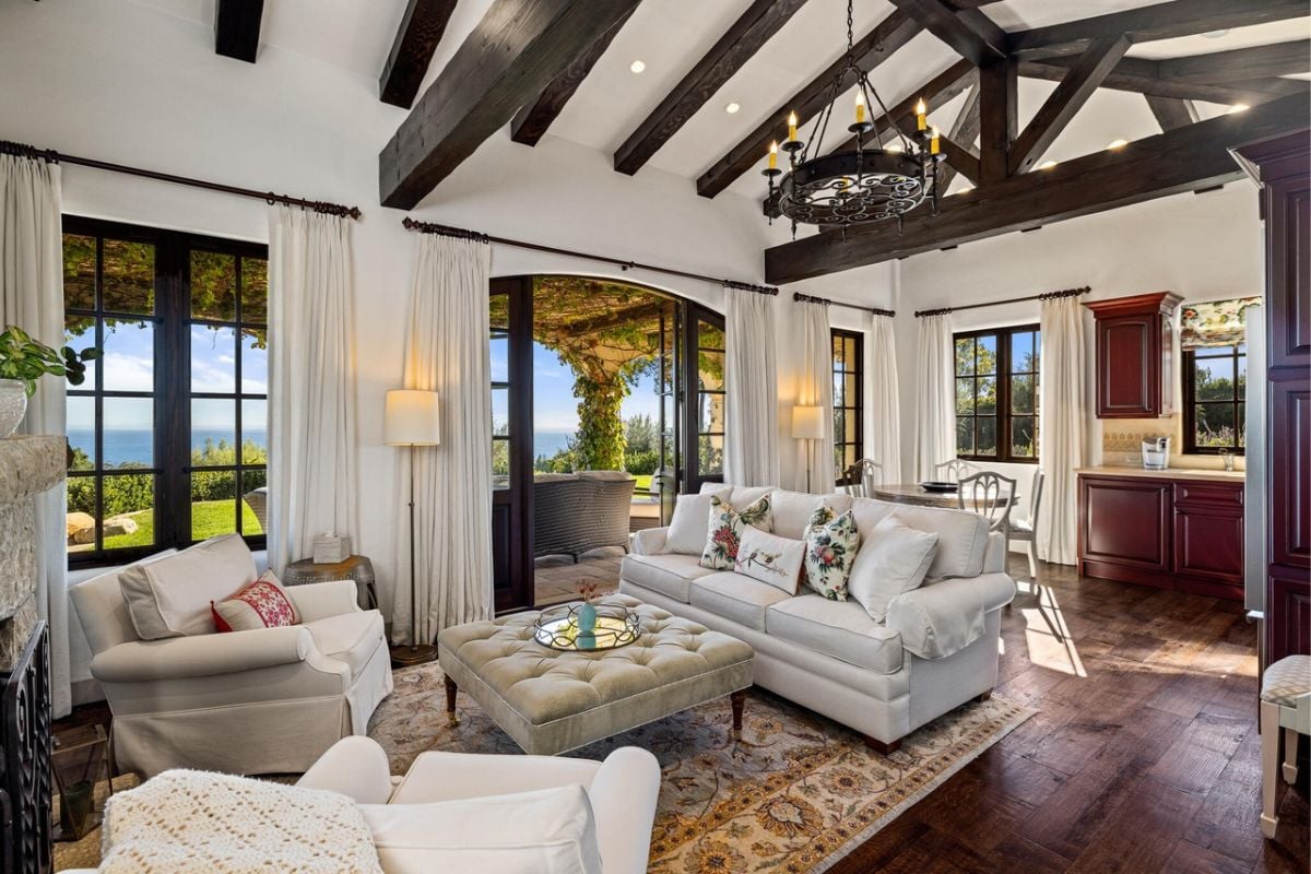 An informal living room with a coffee table surrounded by white couches, a rug, hardwood floor, and a beam ceiling.