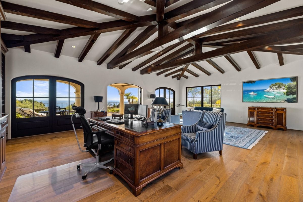 A home office with a wooden desk, chairs, hardwood floor, and open beam ceiling.