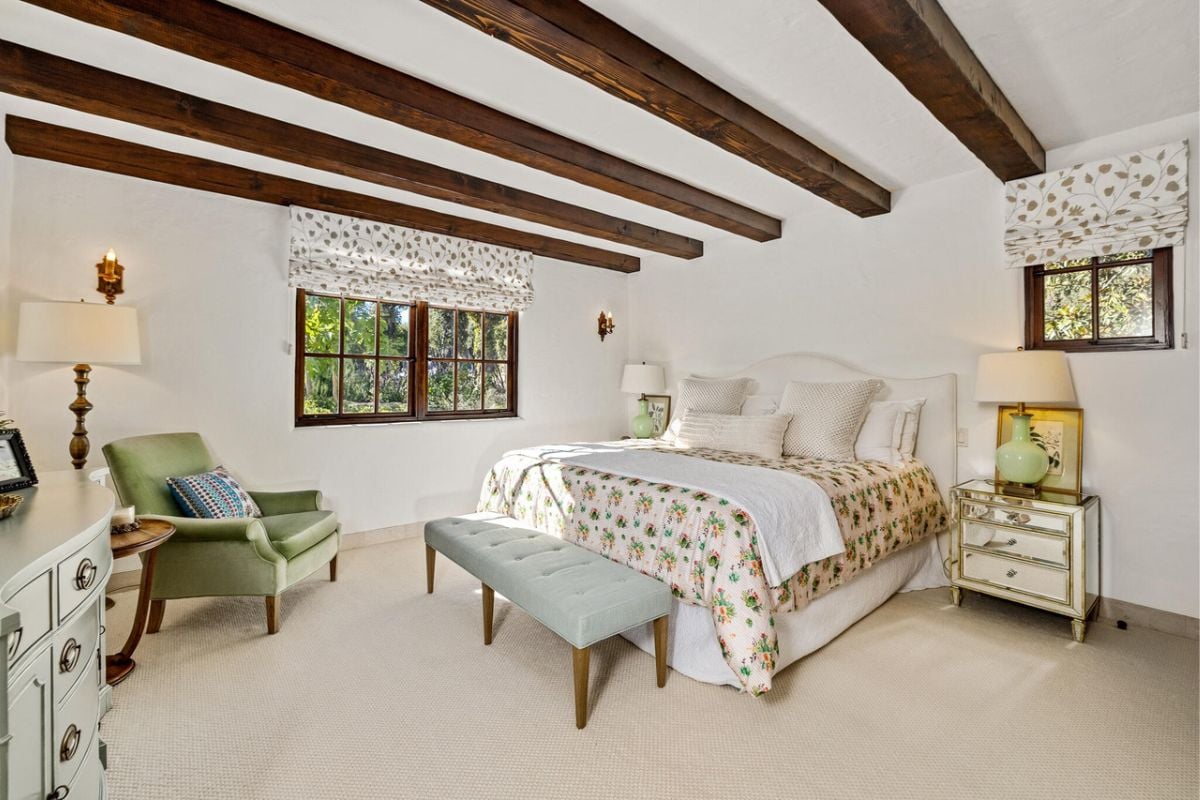 A bedroom with a bed, open beam ceiling, chairs, lampshade, cabinetry, and windows.