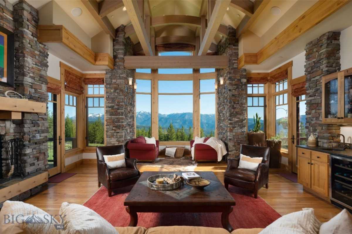 Living room with fireplace, stone walls, exposed beam ceiling, windows, wooden floor, rug, couches, center table, and cabinets.