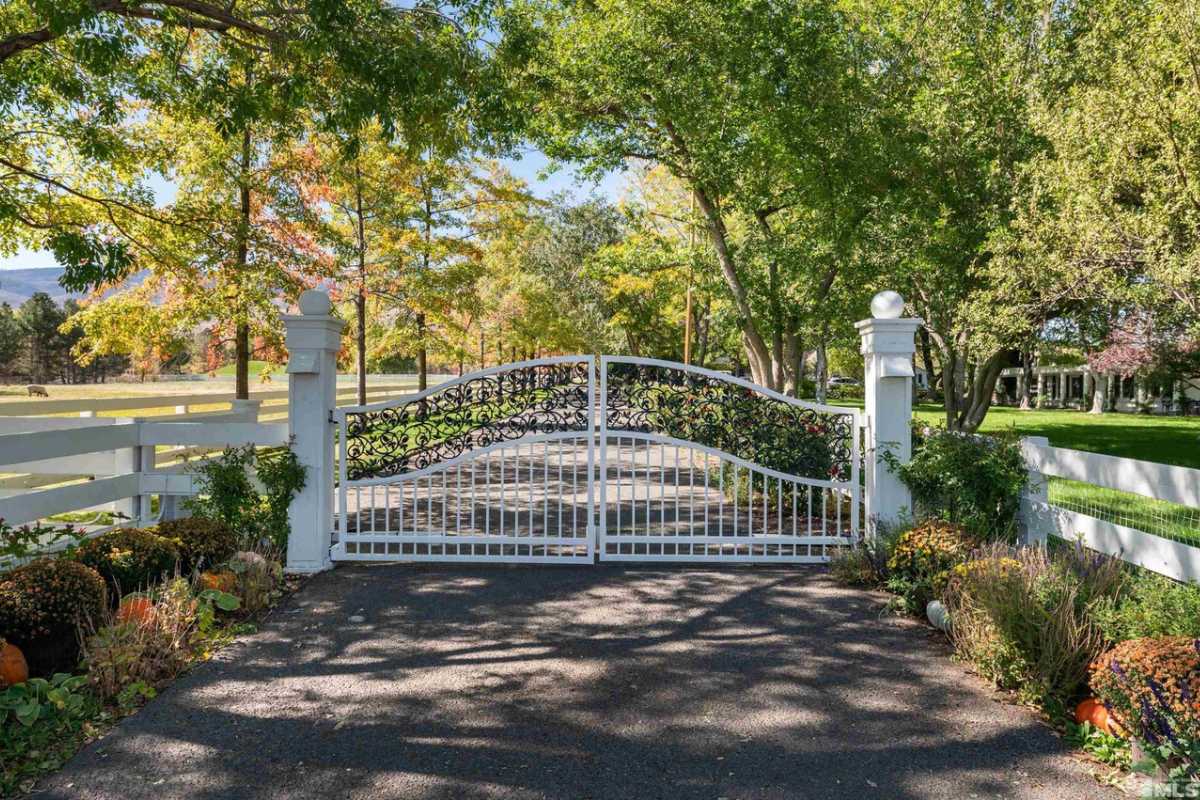 A front gate entry adorned with plants, trees, and a white metal gate.