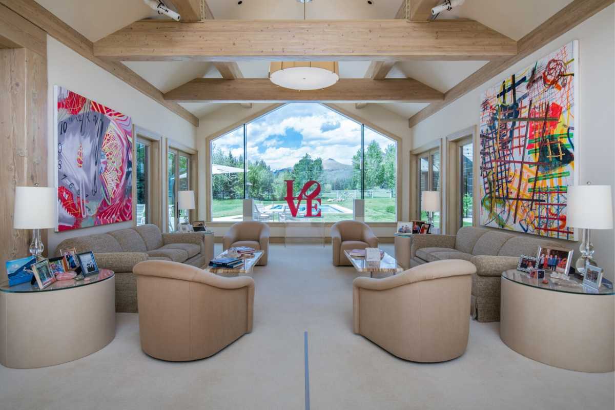 A living room furnished with couches, chairs, a coffee table, paintings, windows, a chandelier, and a beamed ceiling.