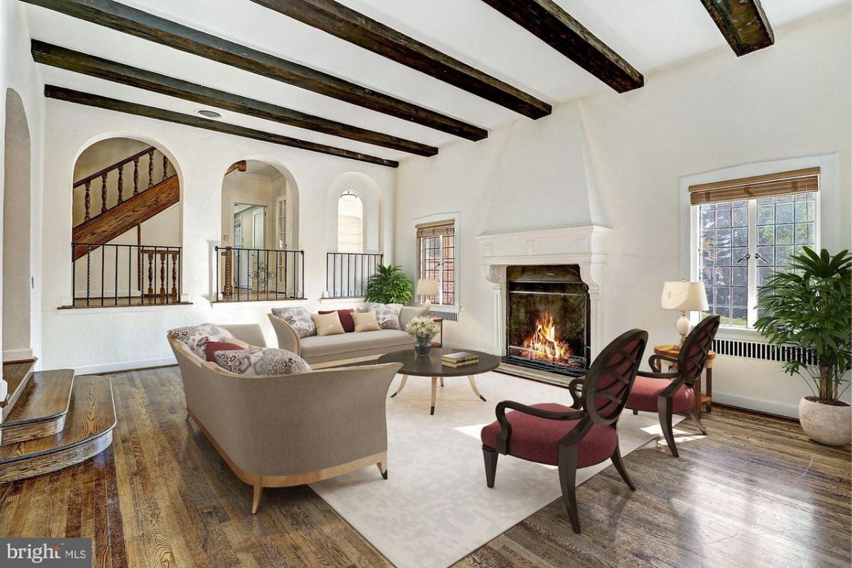 A living room with a wooden floor, wooden beam ceiling, fireplace, plants, and a center table surrounded by couches and chairs.