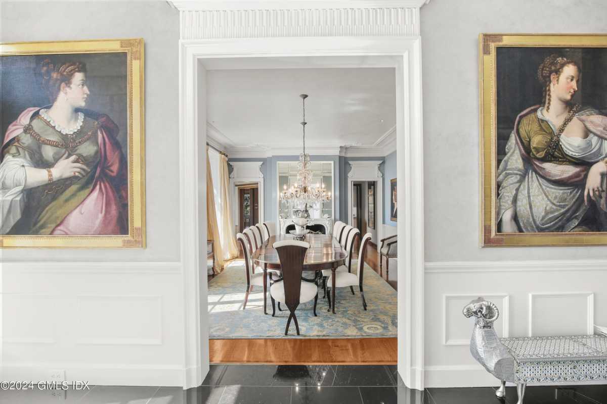 A dining room with paintings, a wooden floor, a rug, a white ceiling, a chandelier, and a dining table surrounded by chairs.