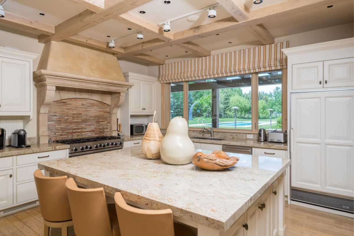 A kitchen with a center island, chairs, sink, countertop, white cabinets, beamed ceiling, windows, and kitchen appliances.