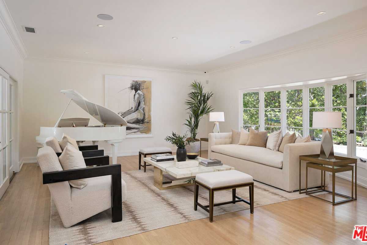 A living room with a wooden floor, windows, a white piano, a white ceiling, a rug, white couches, and a coffee table.