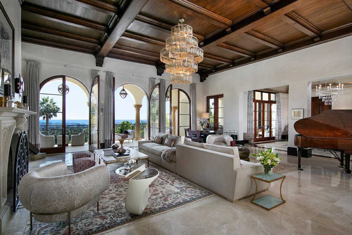 A living room with a tiled floor, windows, a beamed ceiling, a fireplace, a chandelier, a rug, a piano, and couches.