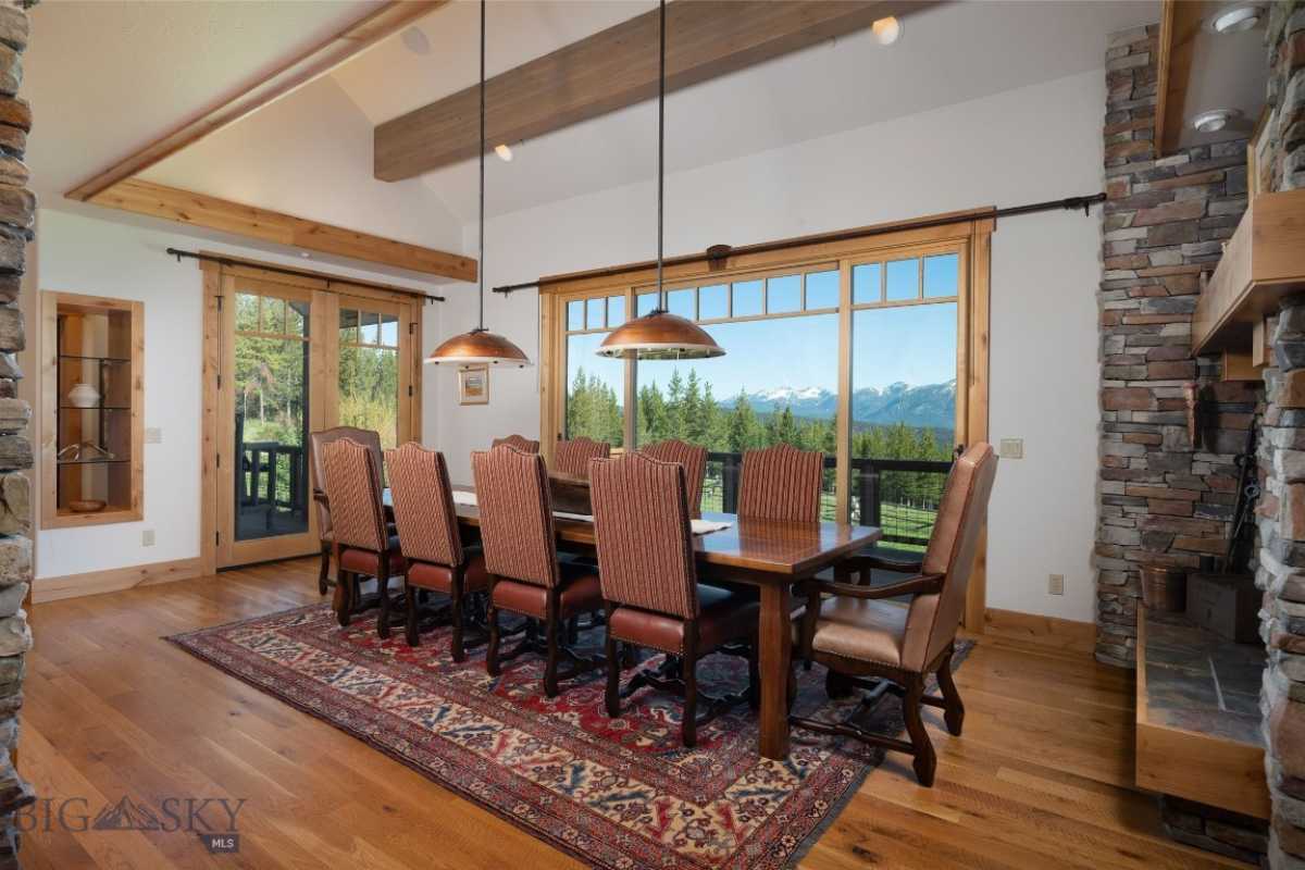 A dining room with a beamed ceiling, window, fireplace, wooden floor, rug, and dining table surrounded by chairs.