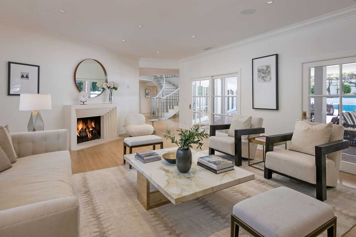 A living room with a fireplace, mirror, wooden floor, windows, a white piano, a white ceiling, a rug, white couches, and a coffee table.