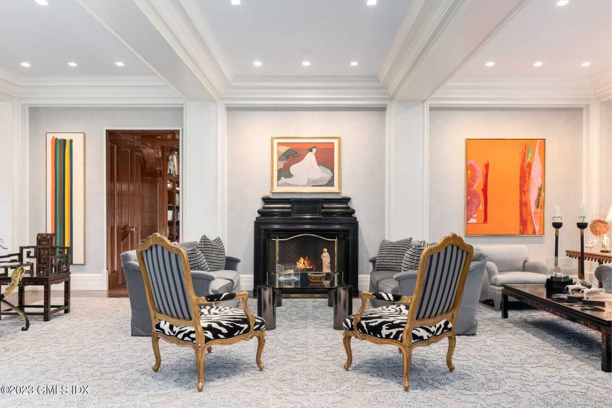 A living room with a fireplace, beamed ceiling, paintings, couch, and chairs.