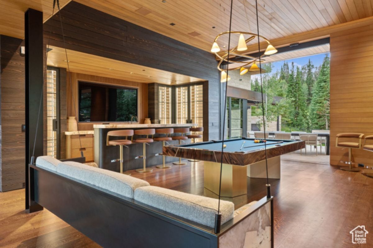 A bar with a wooden floor, wooden ceiling, chandelier, countertop, chairs, couch, and pool table.