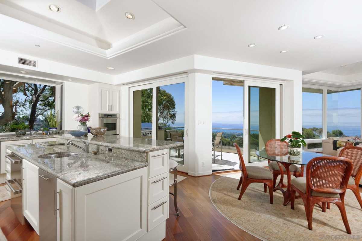 A kitchen with a marble countertop, sink, white ceiling, wooden floor, windows, and a table with chairs.