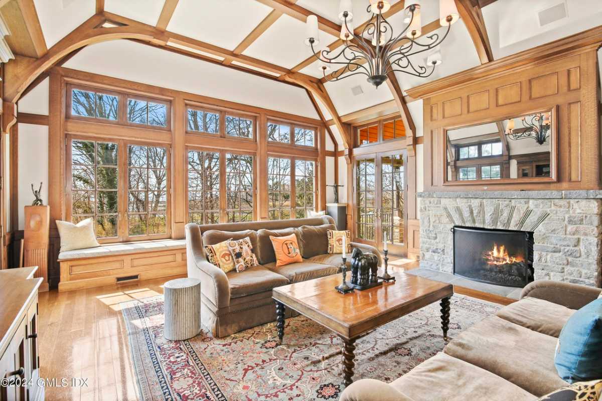 A lounge with a fireplace, wooden floor, windows, exposed beam ceiling, a rug, cabinets, a couch, and a coffee table.