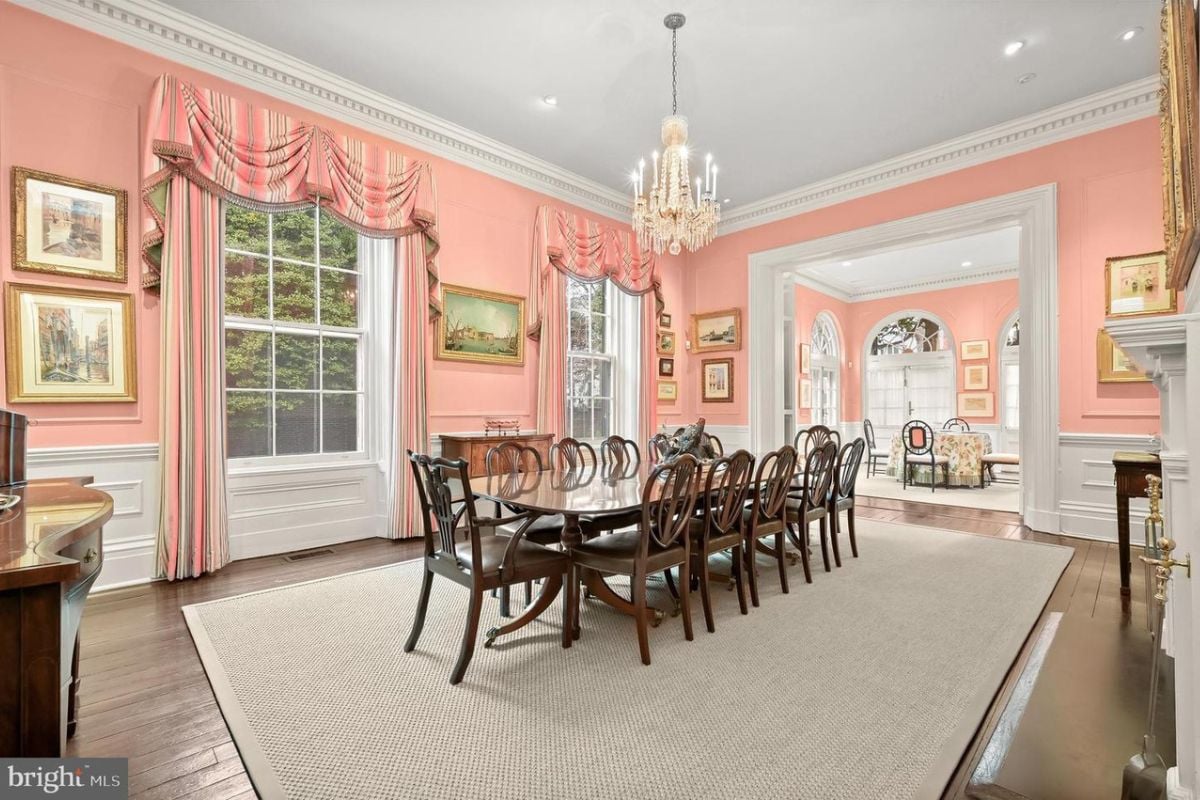A dining room with wooden floor, rug, white ceiling, chandelier, pink walls, fireplace, and a dining table surrounded by chairs.