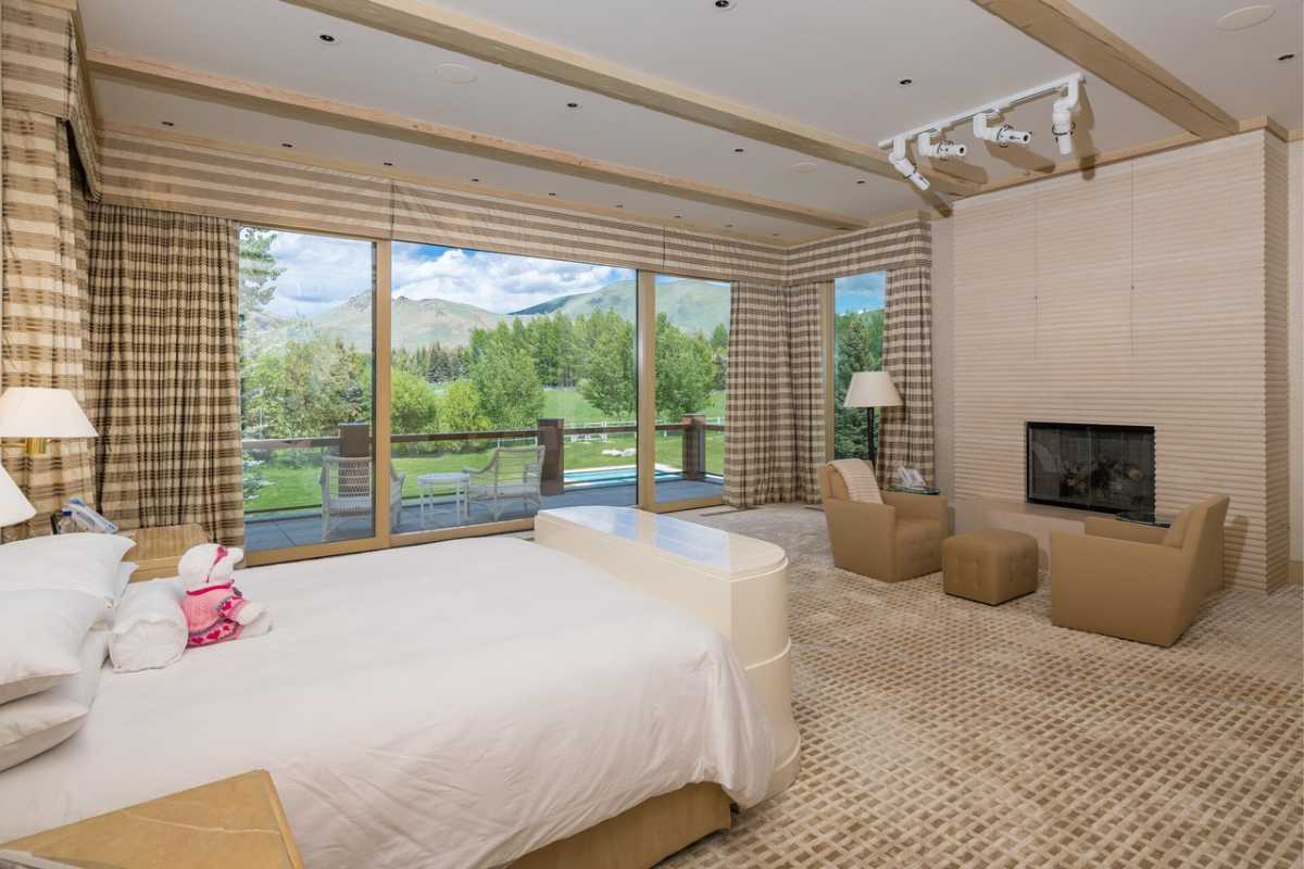 A bedroom with a bed, fireplace, couches, beamed ceiling, and windows.