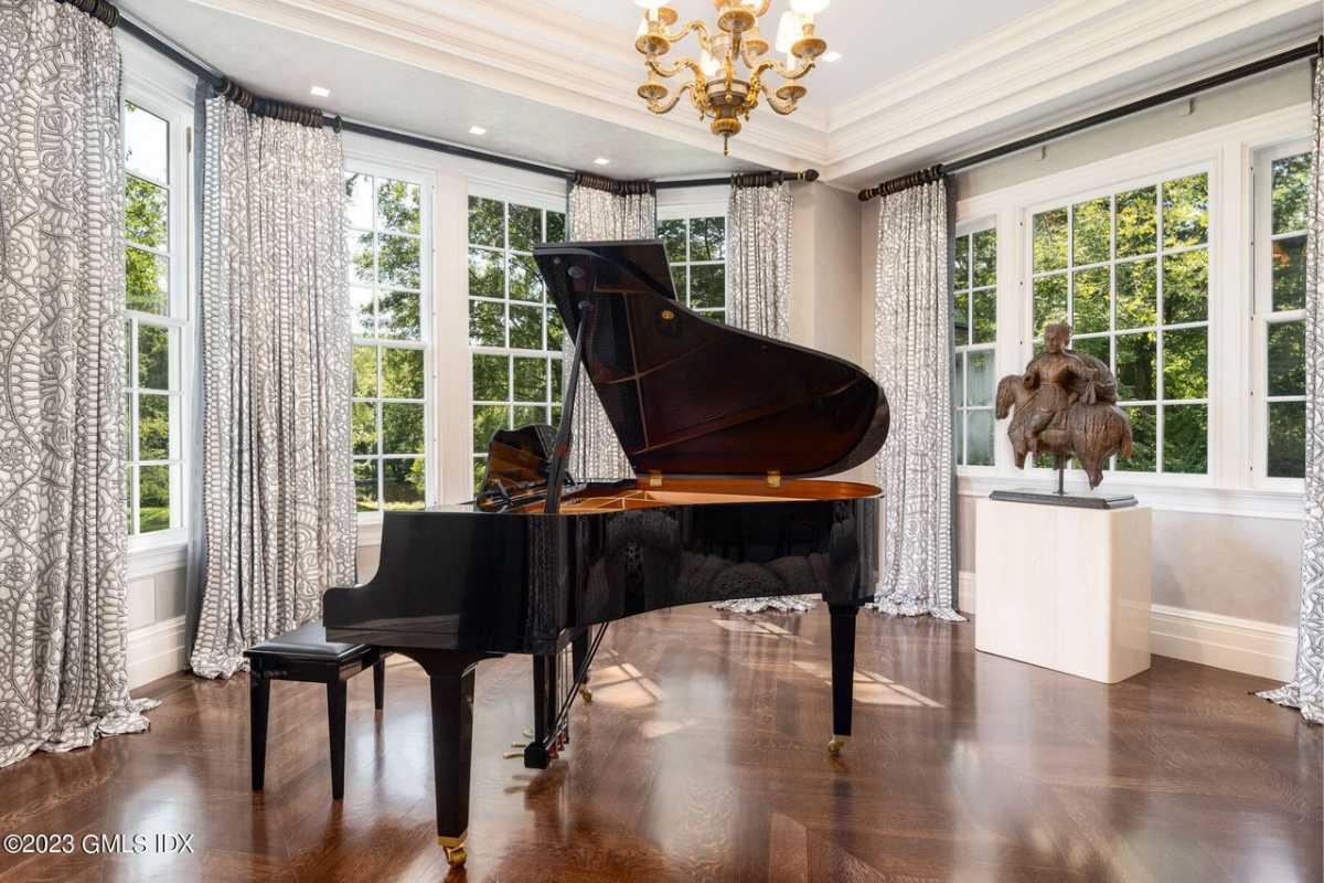 A living room with a piano, wooden floor, chandelier, and windows.