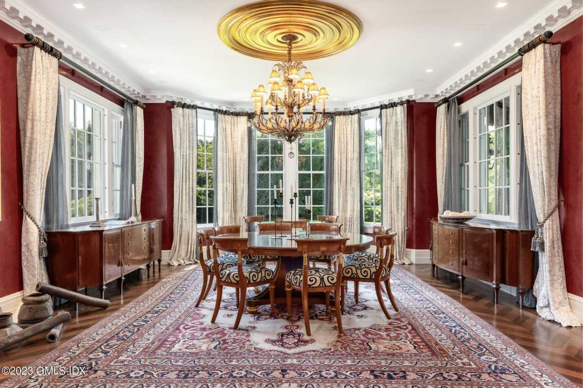 A dining room with a wooden floor, rug, windows, white ceiling, chandelier, and a round table surrounded by chairs.