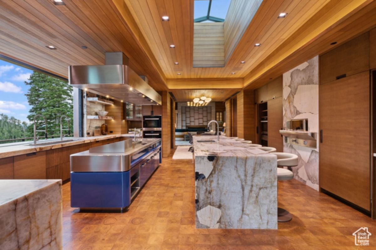 A kitchen with a window, wooden floor, wooden ceiling, marble island, chairs, and kitchen appliances.