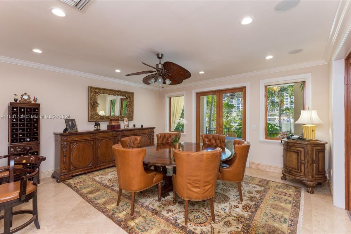 A dining room with a rug, cabinets, lampshade, windows, tiled floor, a white ceiling, a ceiling fan, and a round wooden table surrounded by chairs.