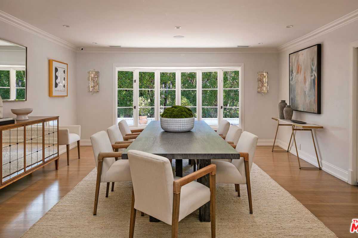 A dining room with a rug, wooden floor, painting, windows, white ceiling, and a dining table surrounded by white chairs.