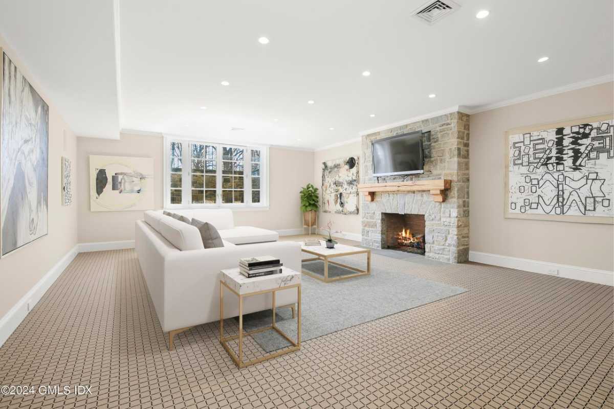 A lounge with a fireplace, paintings, a white ceiling, windows, a TV, a white couch, and a center table.