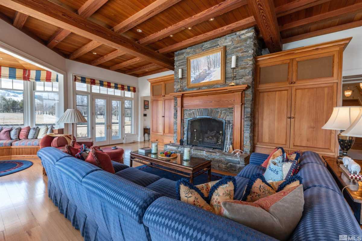 A sunroom surrounded by windows with a blue couch adorned with pillows, a fireplace, wooden floors, cabinets, and an exposed beam ceiling.