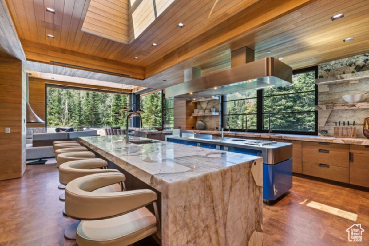 A kitchen with a window, wooden floor, wooden ceiling, marble island, chairs, and kitchen appliances.