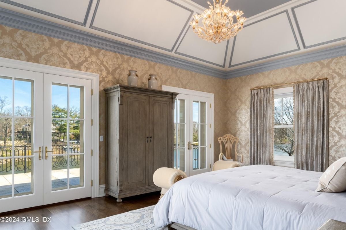 A bedroom with a bed, wallpaper, window, chandelier, chairs, and a cabinet.