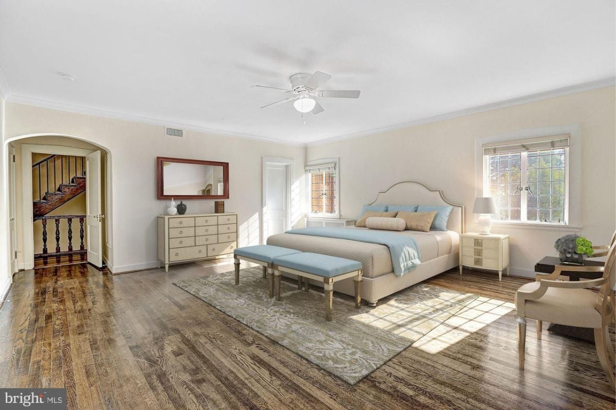 A bedroom with a bed, wooden floor, white ceiling, ceiling fan, windows, nightstand, and lampshade.