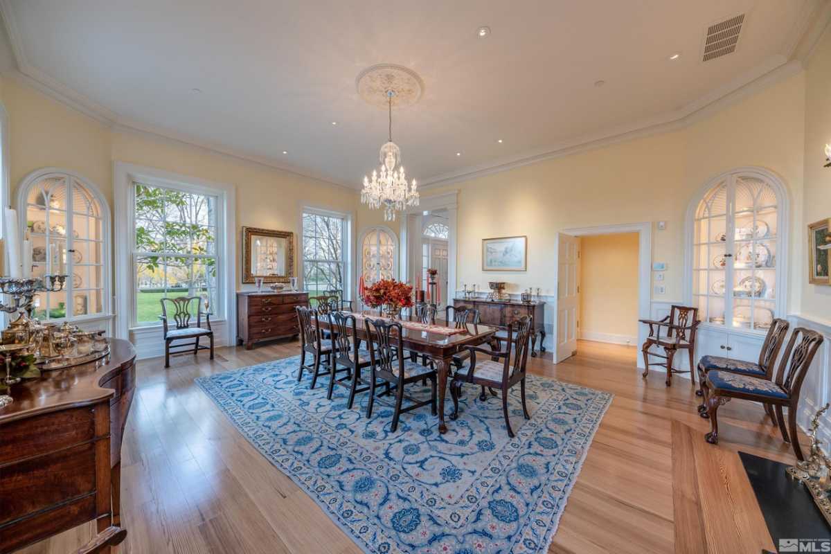 A dining room with wooden floors, a rug, yellow walls, windows, a white ceiling with a chandelier, cabinets, and a dining table surrounded by chairs.